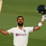 Virat Kohli raising his bat and celebrating after scoring a Test century, wearing the official Indian Test jersey, with the stadium in the background.