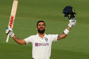 Virat Kohli raising his bat and celebrating after scoring a Test century, wearing the official Indian Test jersey, with the stadium in the background.